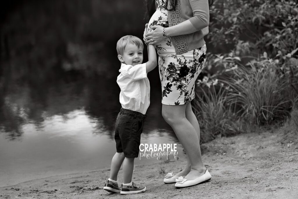 Black and white outdoor maternity photo with toddler smiling at camera while resting hand on mom's baby bump