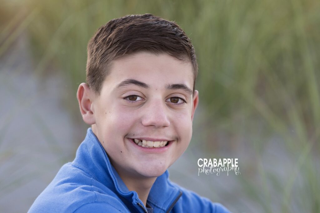 Smiling boy in blue shirt in grassy dunes during coastal family session