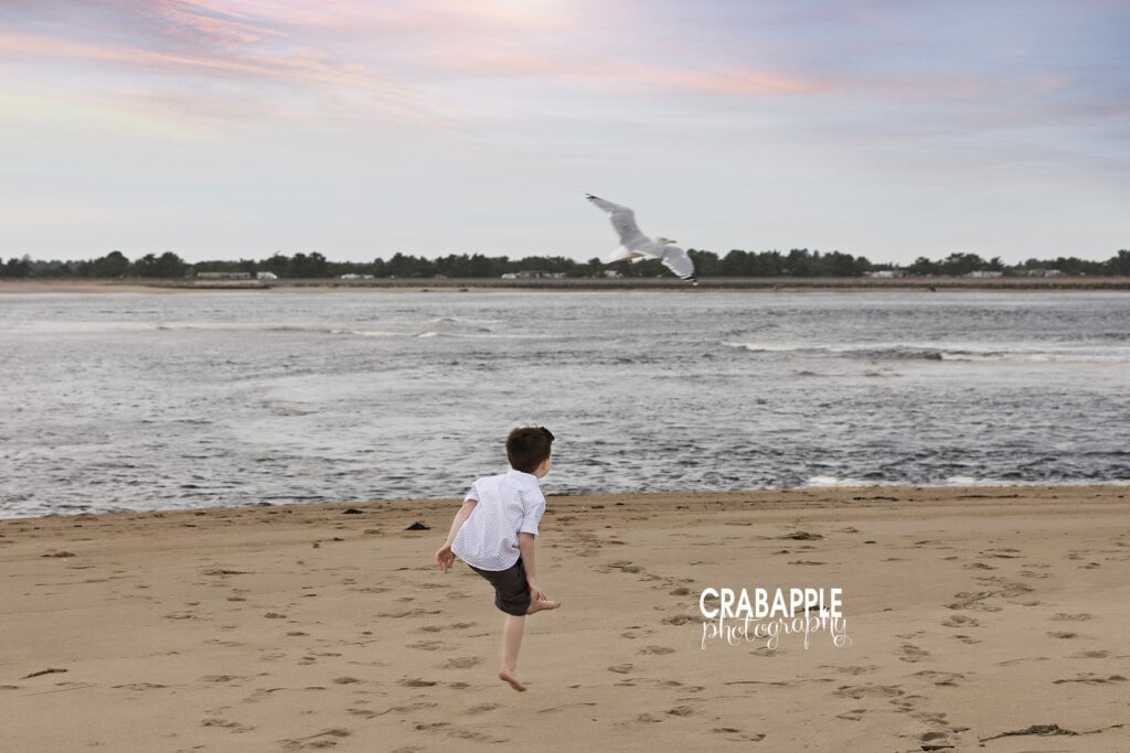 Toddler gazing out at ocean waves and spotting a seagull during Salisbury Beach photo session a fun example of childhood at the beach