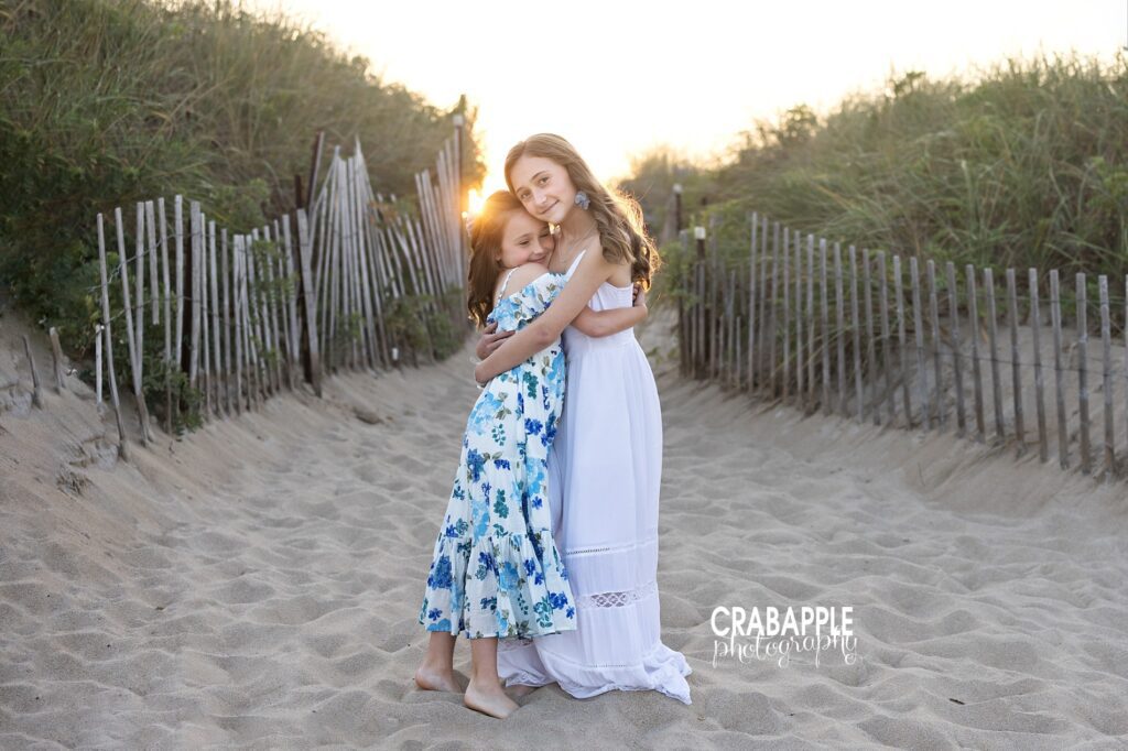 Boston beach photo of sisters with golden hour sun flare Older sister hugging little sister with golden sun flare at Boston beach during golden hour photography session