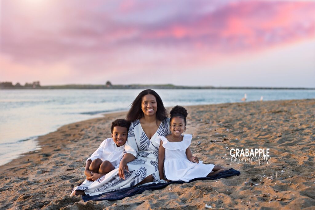 Mom and two children sit on a beach blanket in the sand with gorgeous sunset views behind them during a mini session