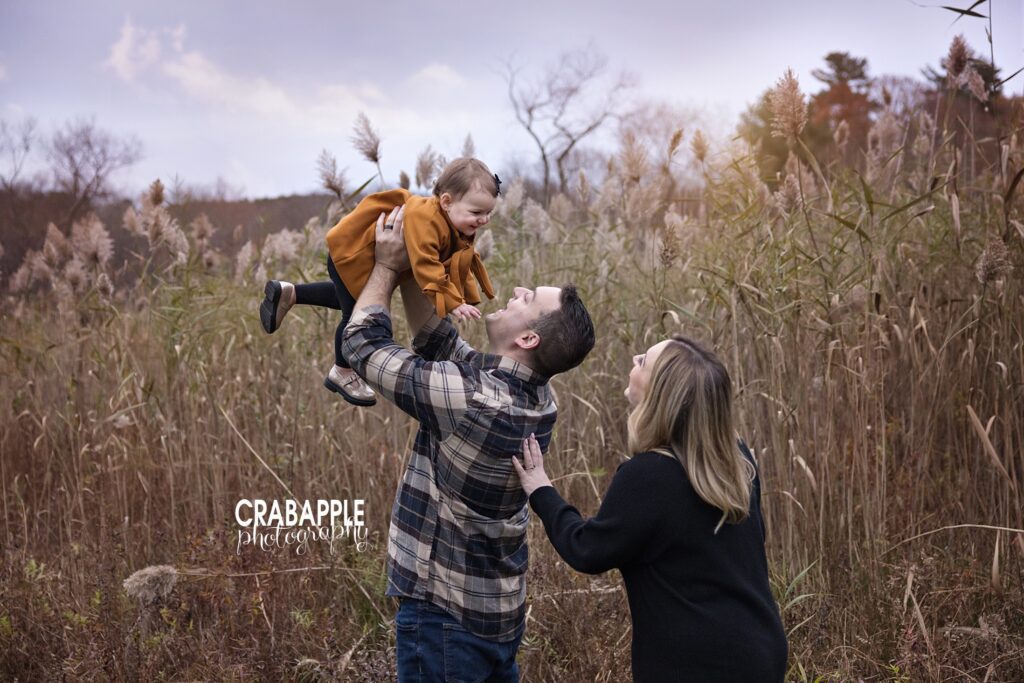 Fall baby photos with goldenrod outfit Baby girl in mustard goldenrod coat at fall photo session with dad lifting her into the air as mom watches