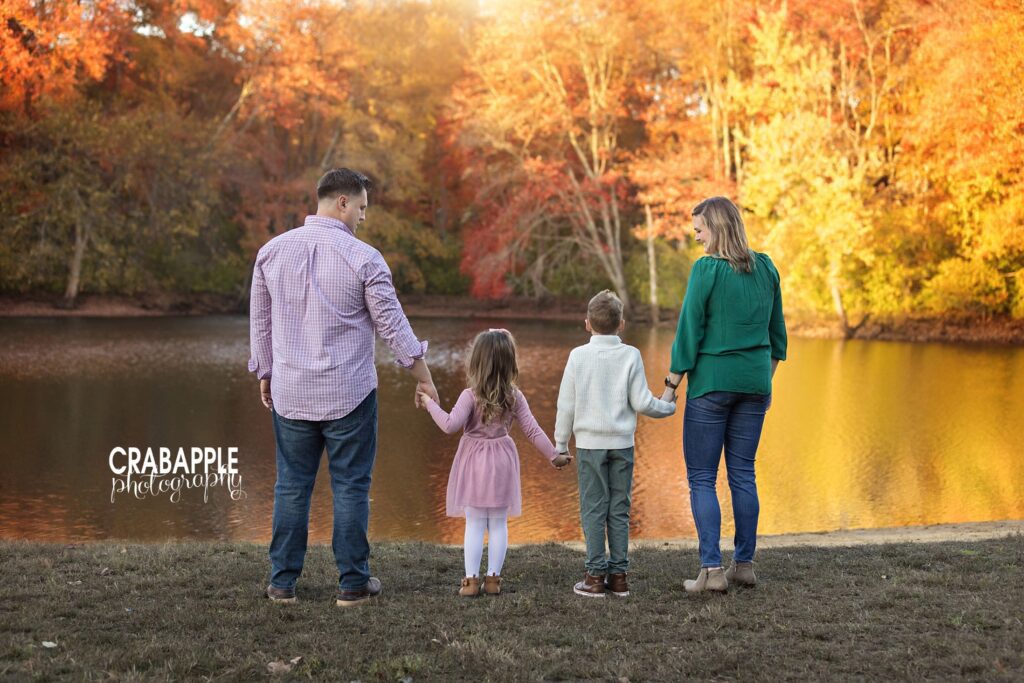 Family of four face away from the camera and toward a pond surrounded by fall foliage in candid family portrait