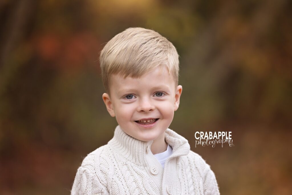 Outdoor fall child portrait in Lynnfield MA of 4-year-old boy in cream sweater surrounded by autumn leaves
