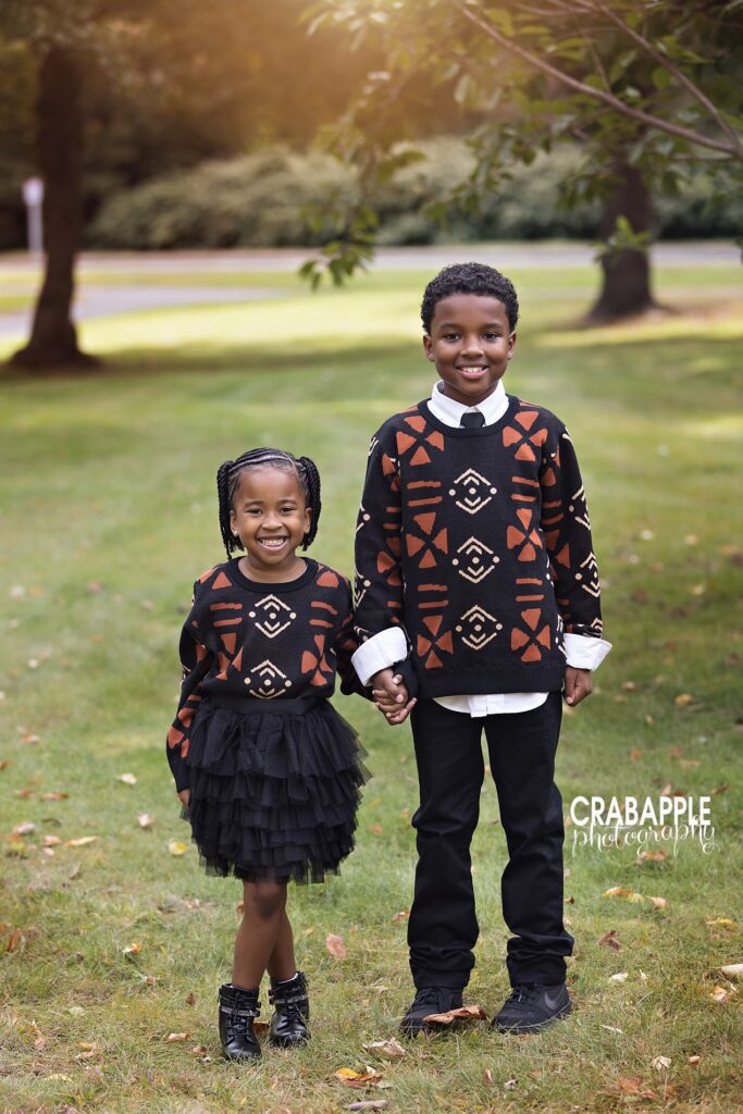 Two siblings, older brother and younger sister, hold hands and smile at the camera in matching outfits