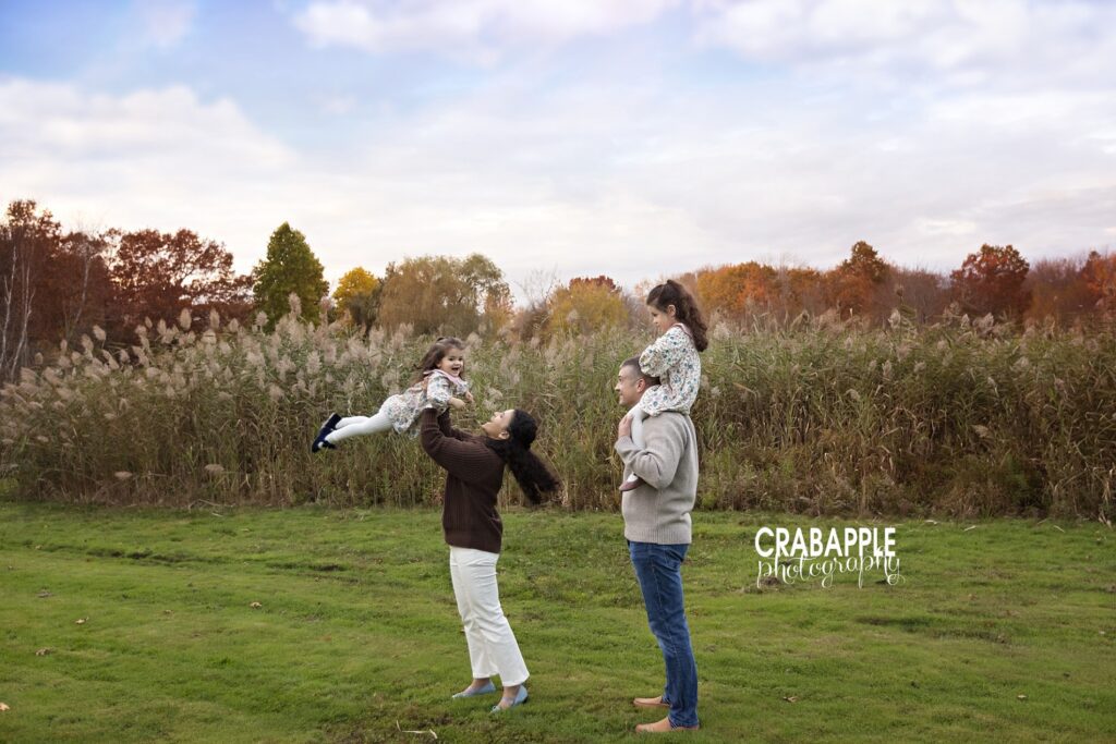 Dad carrying daughter on shoulders while mom swings other daughter in the air in Andover fall family portraits Dad carries his daughter on his shoulders and watches while mom swings younger daughter in the air during fall family portraits in Andover Massachusetts