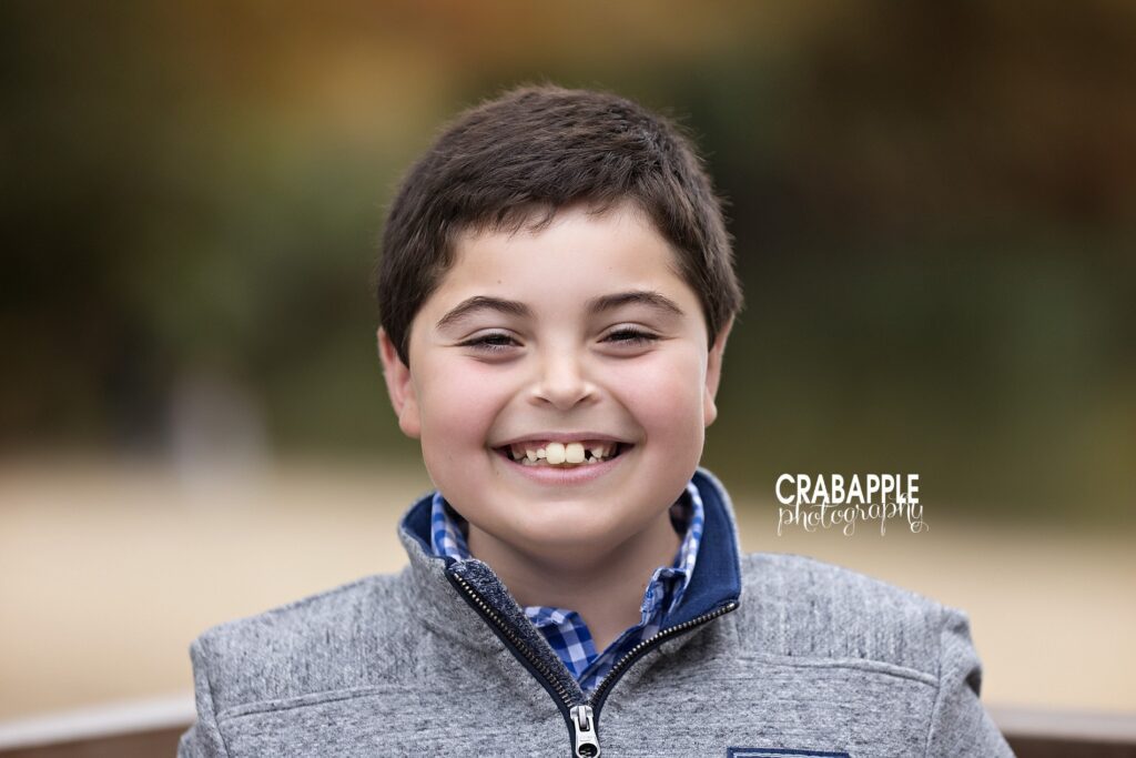 A boy smiles while wearing a gray sweater over a blue and white plaid shirt during fall family photos 