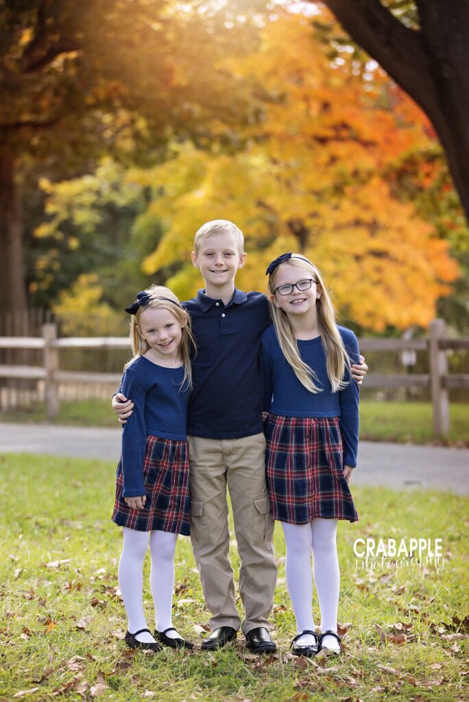 Three siblings in coordinated outfits stand in front of fall foliage during family portrait session in North Shore MA