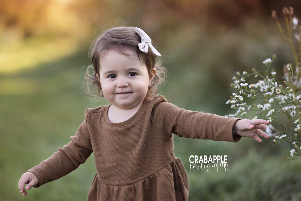 Toddler girl wearing a brown outfit with bow in hair smiles at camera during Andover MA area family portrait session