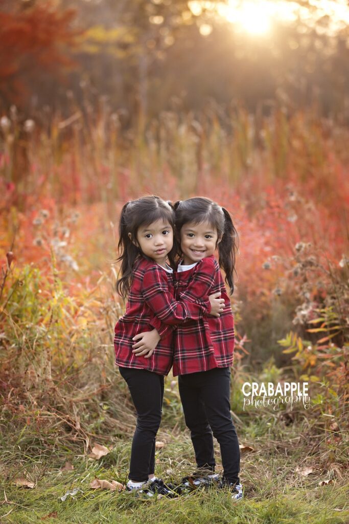 Twin sisters hug one another during family outdoor fall photo session with foliage and sunset behind them