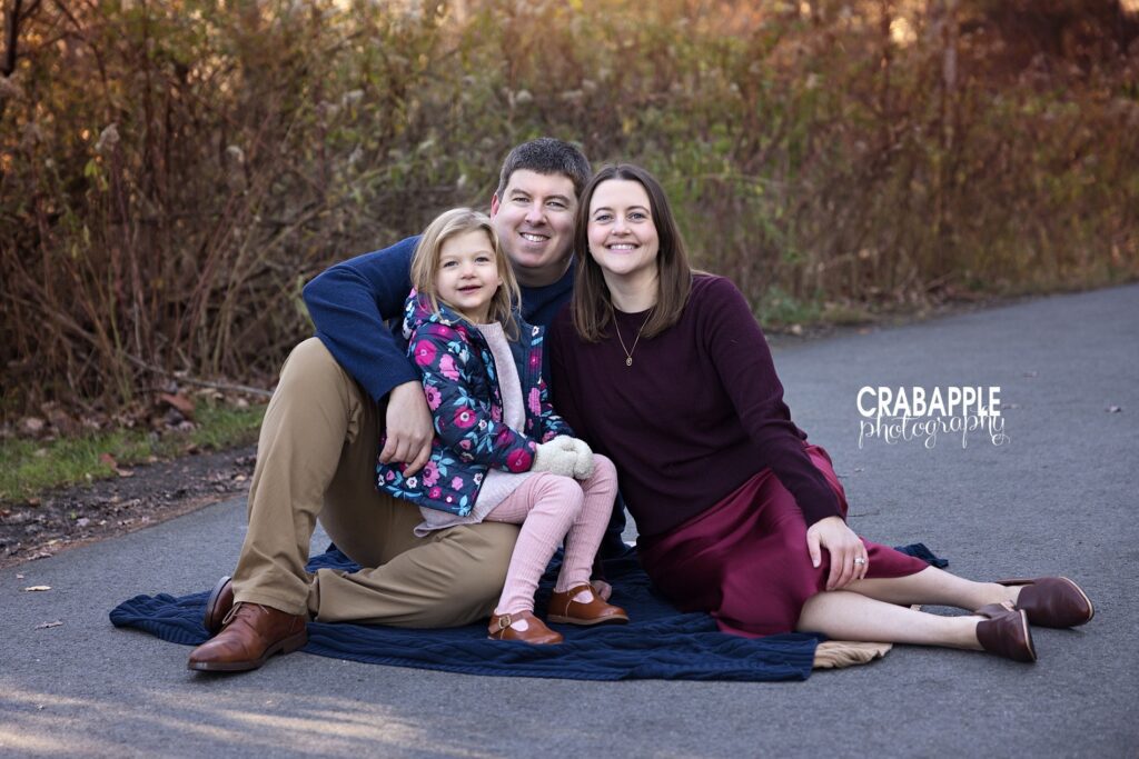Waltham MA family portraits outside Family of three smile together on a blanket on the ground during an outdoor family portrait session near Waltham MA