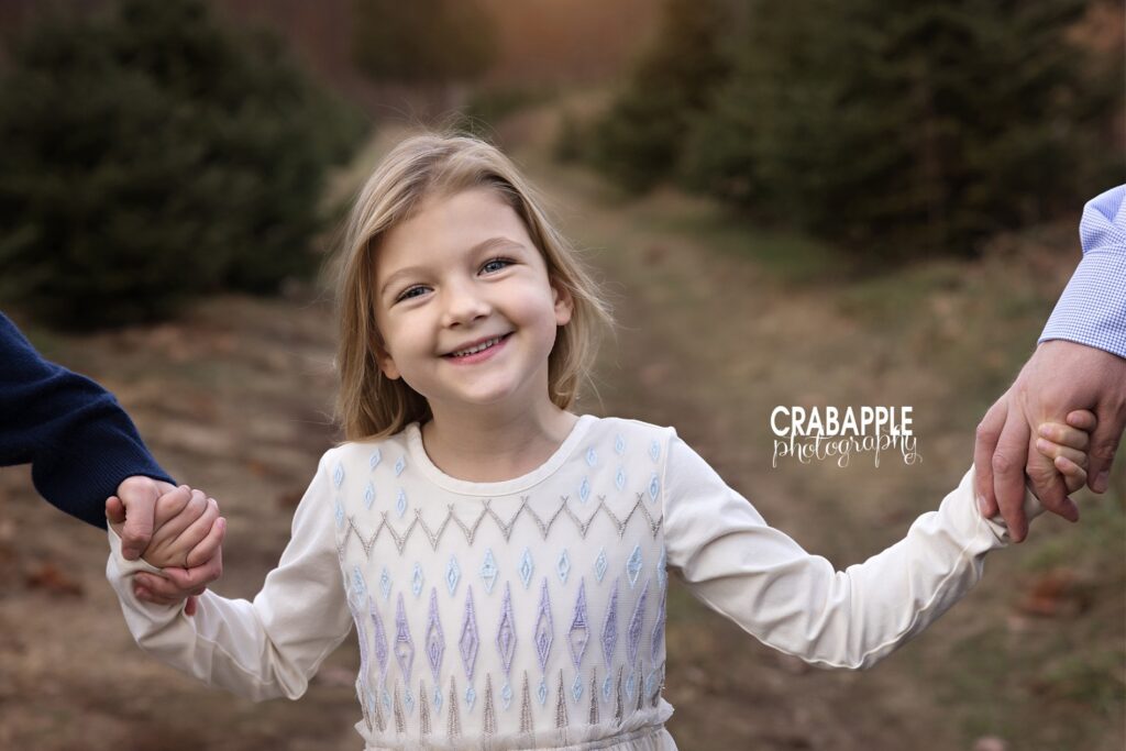 A young girl holds hands with her parents, who are off camera, during family tree farm session