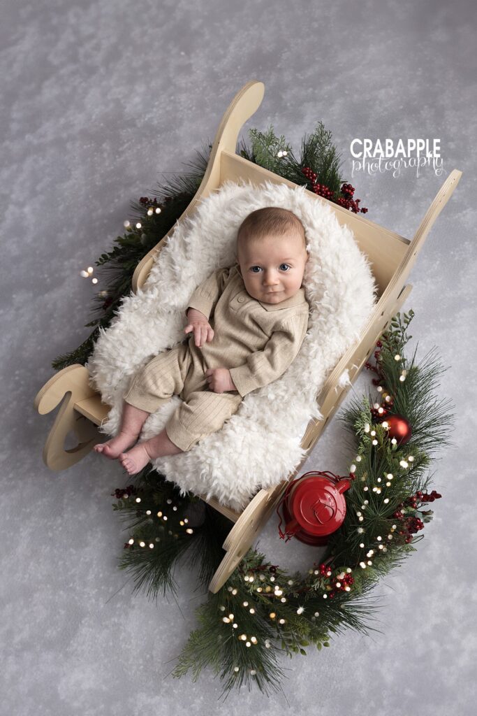 Cozy holiday portraits for infants | Christmas baby sleigh photo Infant peacefully resting in wooden sleigh prop and surrounded by pine bough, twinkling lights and ornaments for cozy holiday portraits for infants.