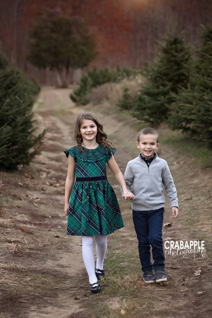 Siblings in coordinating holiday outfits walking hand in hand at tree farm
