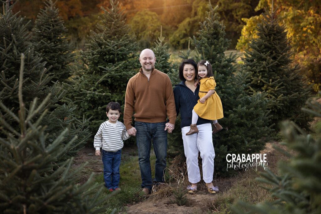 Family of four pose in front of pine trees in a mix of neutral and vibrant outfits for holiday photos