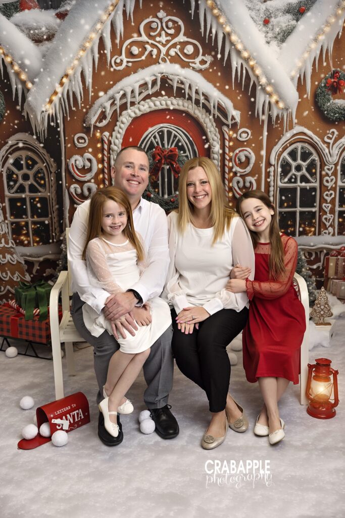 Mom, dad, and two daughters sitting in front of a gingerbread house backdrop during family Christmas session.
