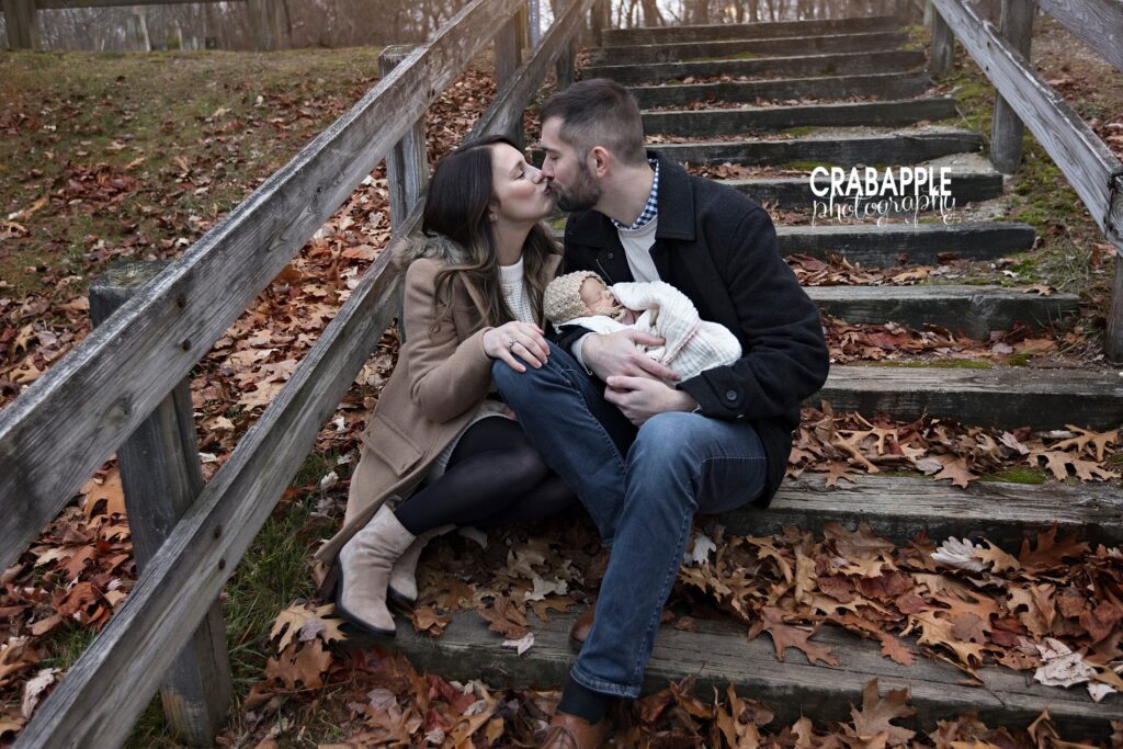 Parents sharing a kiss while holding their baby on rustic wooden steps surrounded by fallen leaves
