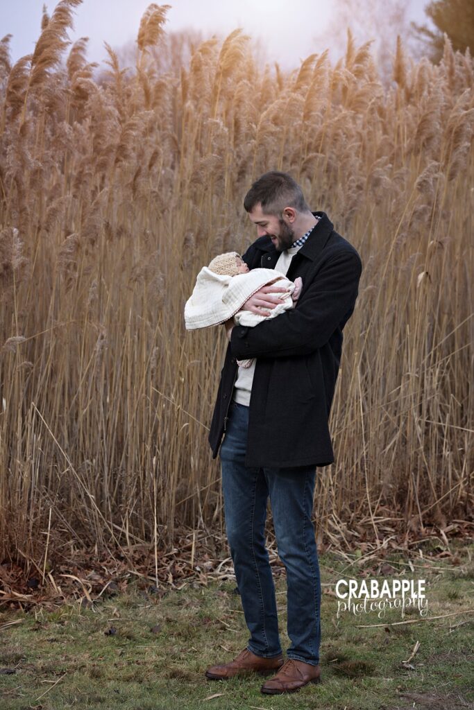 Dad holding newborn baby wrapped in a cream blanket in front of tall grasses
