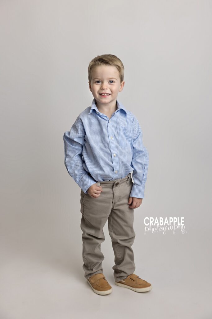 Toddler and child portrait ideas Groveland MA Portrait of a young boy in blue button up standing in front of a simple white backdrop and smiling