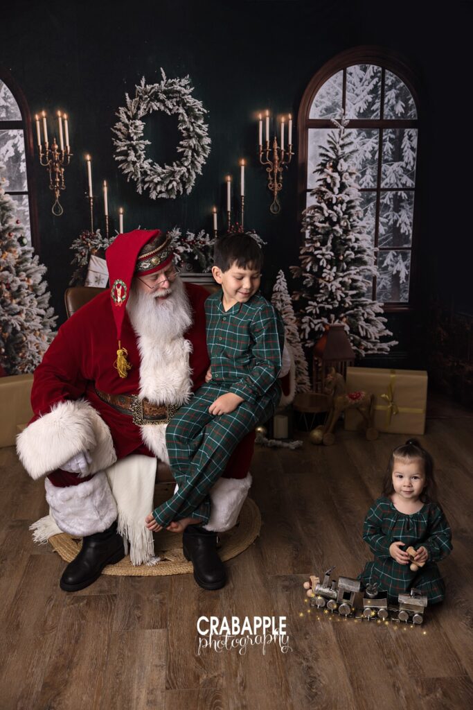 A young boy sits on Santa's lap while his toddler sister plays with toys in front of them