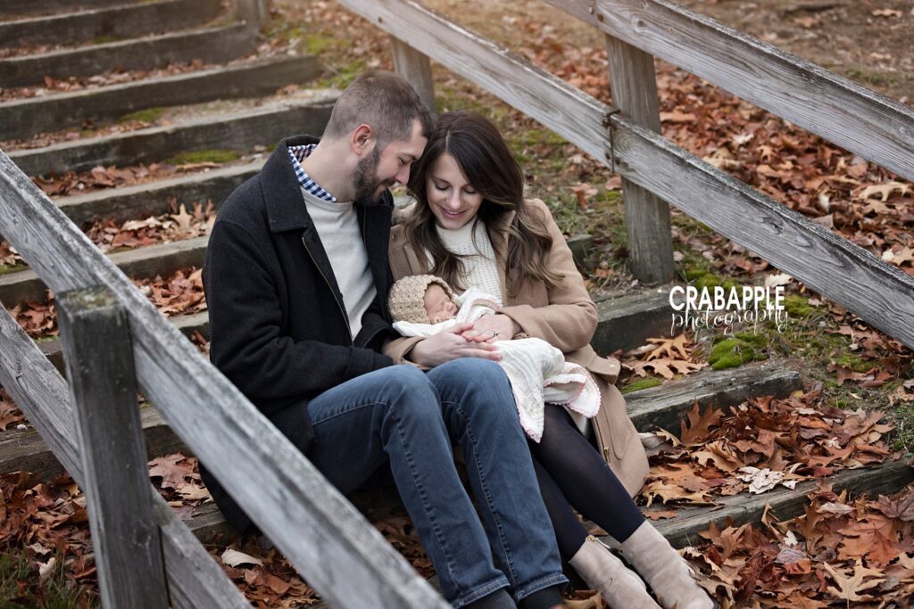 Mom and dad smiling down at baby wrapped in a neutral knit blanket outdoors
