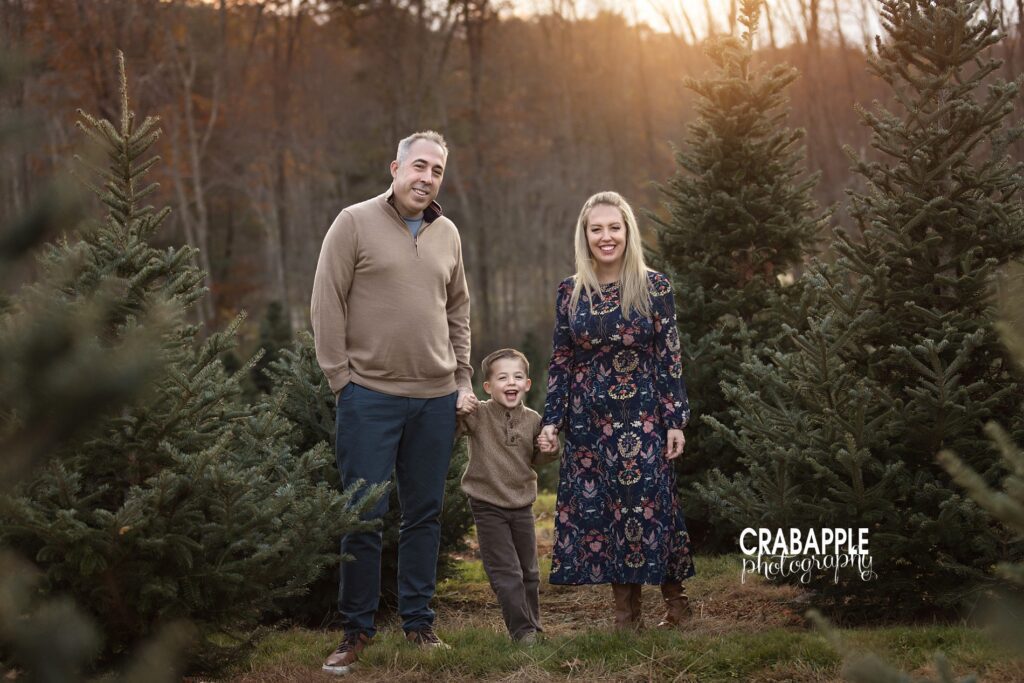Mom and dad hold hands with son as they pose in front of golden sunlight and pine trees
