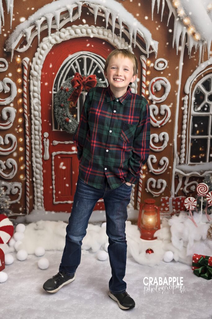 A young boy in a plaid shirt smiles for the camera in front of a gingerbread house backdrop during Christmas photos in Andover MA