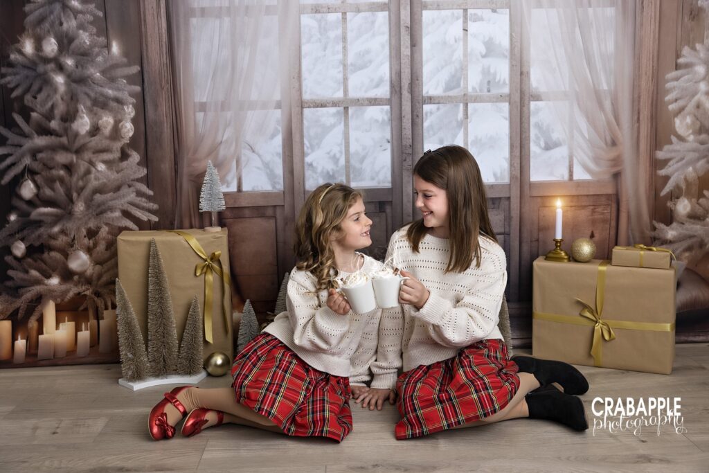Two sisters clink mugs of hot cocoa together during their family and child Christmas portrait session