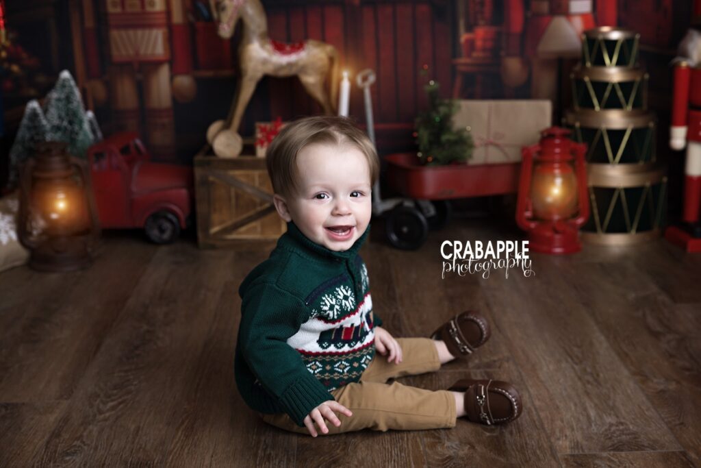 1 year old boy in green sweater and khakis sitting on festive holiday backdrop smiling at camera
