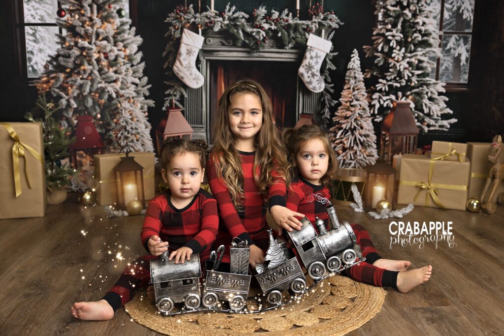 Three sisters in matching plaid pajamas posing with silver Christmas train decor during festive studio portraits