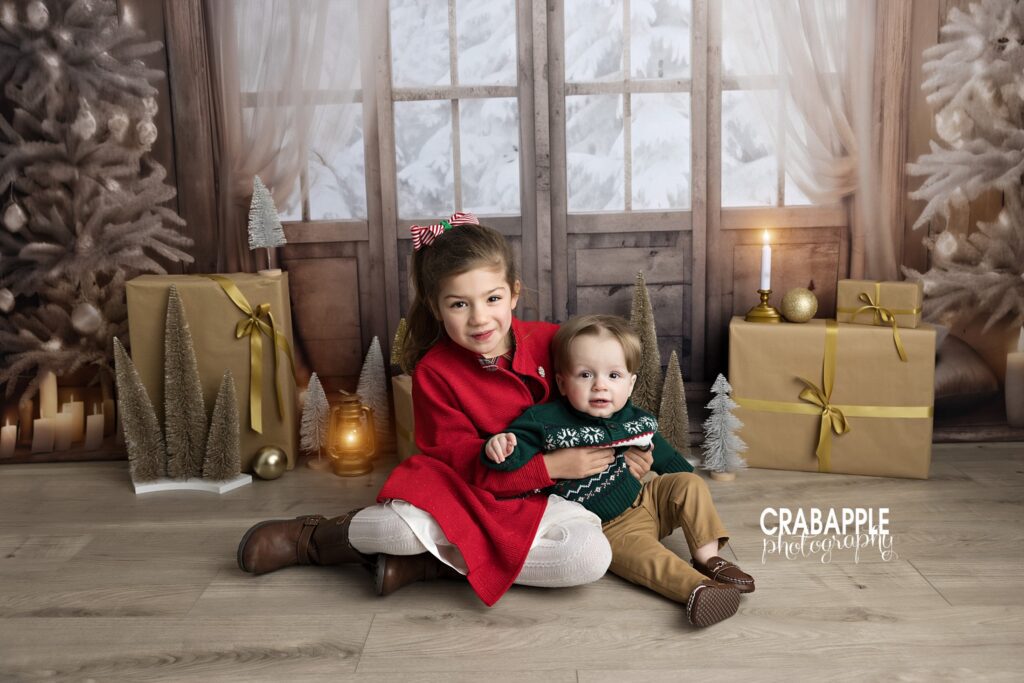 Portrait of 5 year old girl and 1 year old boy sitting together in front of a beautiful and classic Christmas set design in a Haverhill MA area photography studio