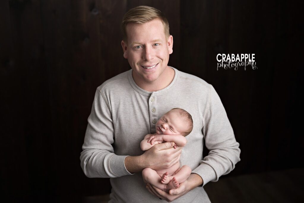 Father smiling while holding his swaddled newborn against a dark studio backdrop.