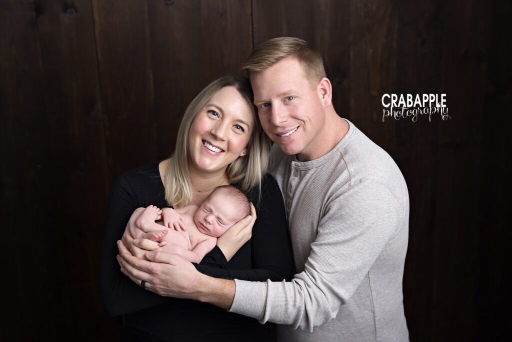 Family of three smiling while holding newborn baby wrapped in gray swaddle.

