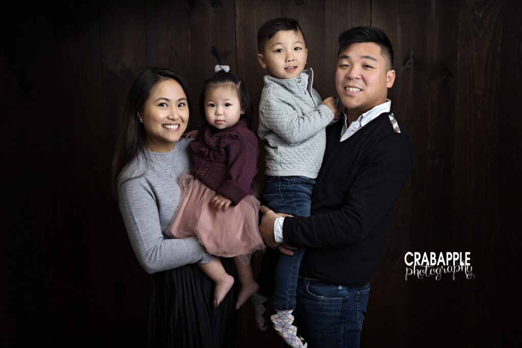 Family of four smiling together during Quincy MA first birthday photos with dark wood backdrop.
