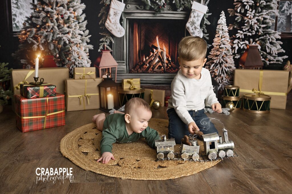Two brothers playing with a toy train in front of a cozy Christmas fireplace set.

