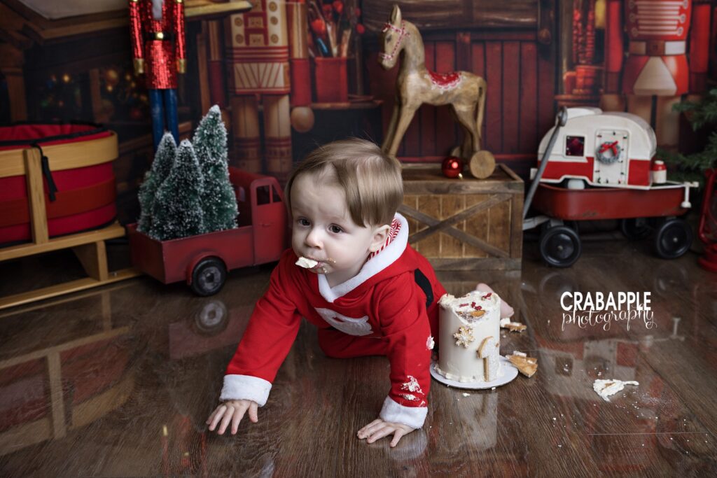 A funny photo of 1 year old boy in Santa outfit crawling away from his cake with cake in his mouth during a Christmas cake smash session