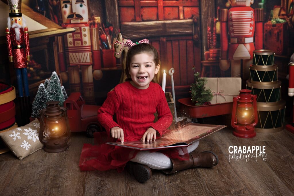 1 5 year old girl in red outfit smiles for the camera while reading a book on a festively designed photography studio set for the holidays