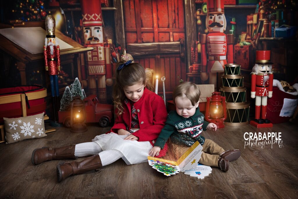 Two cousins (5 year old girl and 1 year old boy) read a book together on a festive photography studio set during a holiday portrait session