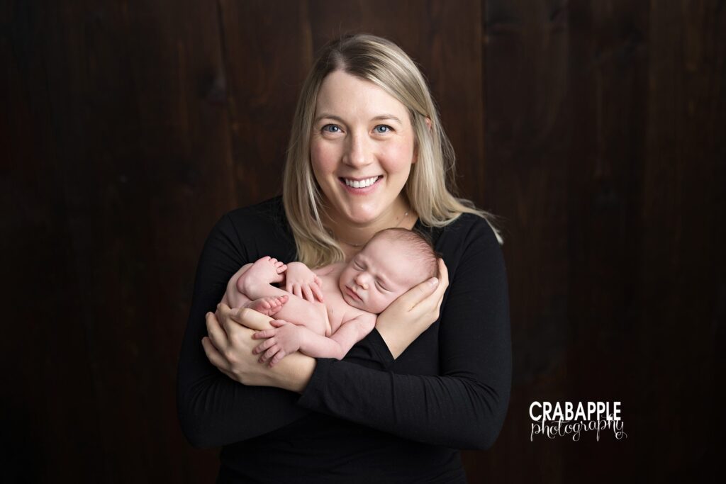Mother smiles while holding her newborn baby close to her chest against a dark backdrop during Boston area newborn portraits