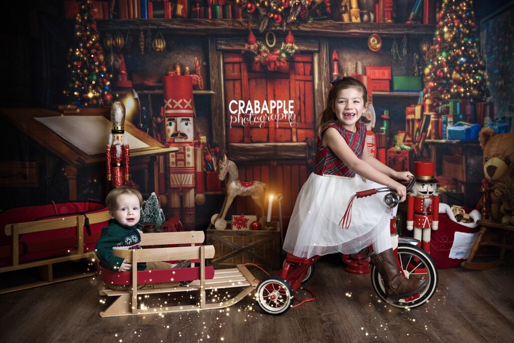 Two cousins on a festive holiday themed set at Andover MA photography studio. Older cousin (5 year old girl) rides a tricycle and pulls younger cousin (1 year old boy) in a sleigh.