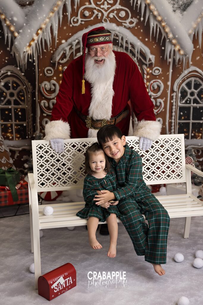 Pajama siblings with Santa for Santa photo gifts Brother and sister in matching pajamas sitting on a bench with Santa behind them, ideal for Santa photo gifts and holiday portrait gift ideas.