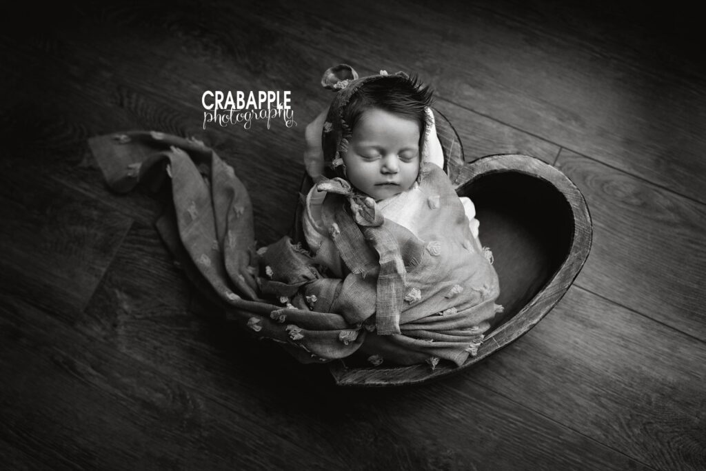 Black and white newborn portrait of a baby girl asleep in a heart shaped wooden bowl on a dark wooden floor