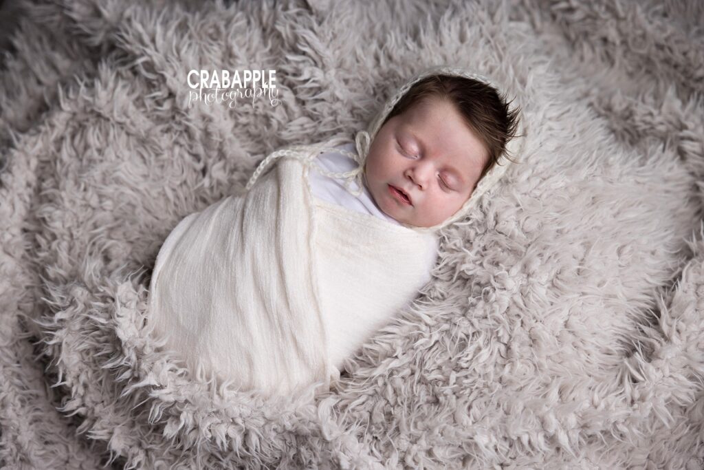 Sleeping baby girl swaddled in white wrap with matching bonnet on a textured off white fur during Ipswich newborn photography session