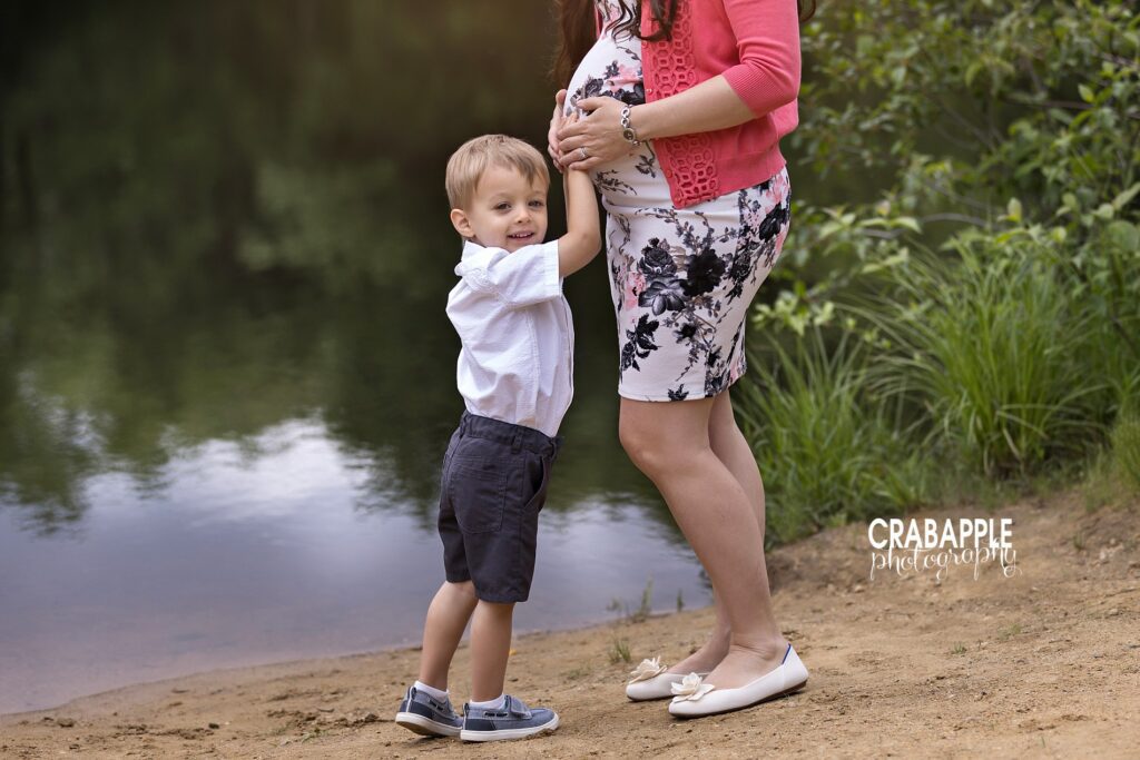 Toddler big brother places hand on mom's baby bump and smiles at camera