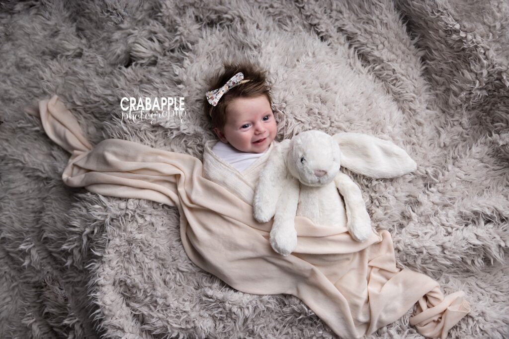 Inspiration for newborn baby girl photos Smiling and awake newborn baby girl on cream colored blanket with a light blush pink wrap and stuffed rabbit
