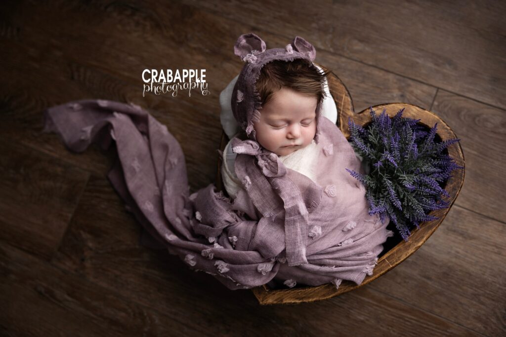 Newborn girl wrapped in light purple with floral accents in a dark wooden heart shaped bowl prop