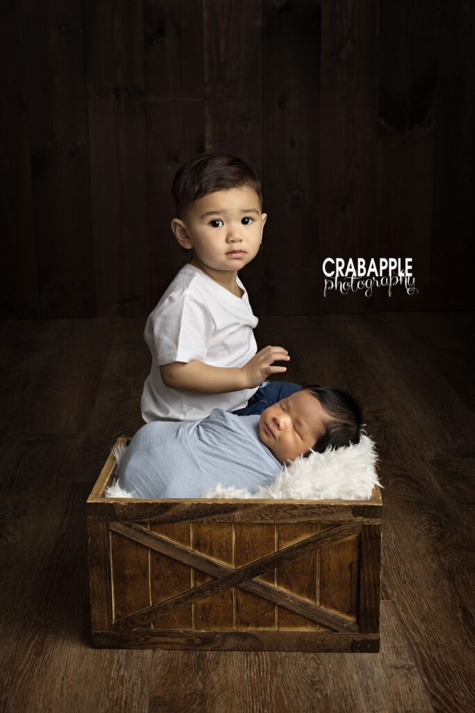 Newborn baby boy posed safely in a wooden box with toddler brother behind him during a studio newborn photoshoot in Quincy MA
