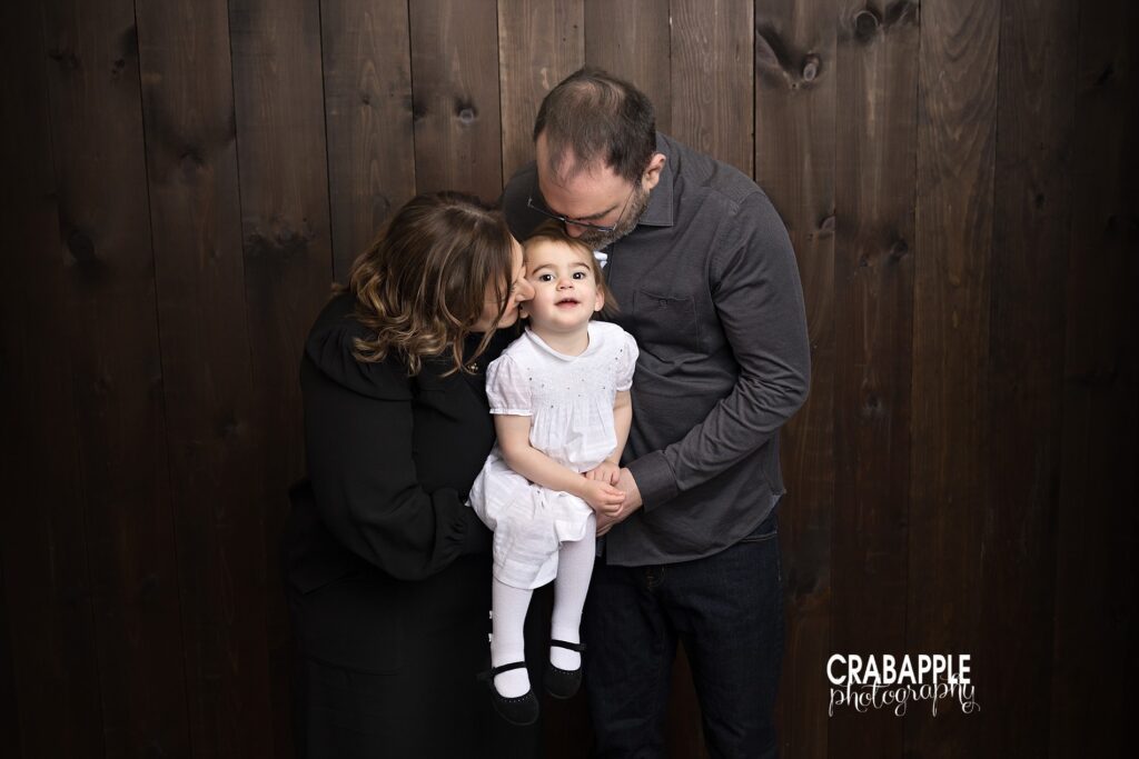 Studio portrait of a two year old girl held by her parents against a dark wood backdrop

