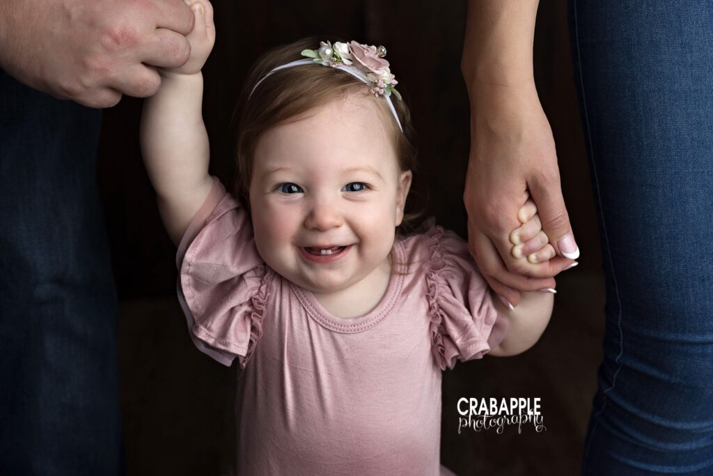 baby girl standing holding parent hands dark backdrop

