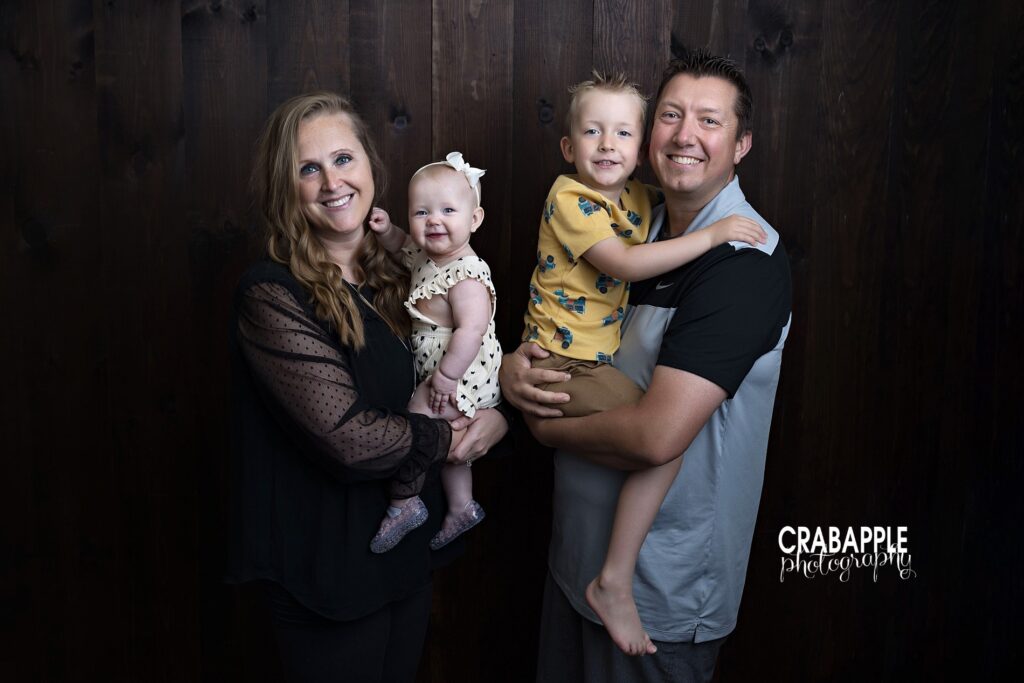 Family of four smiles and poses in front of a dark wooden backdrop in Andover MA studio